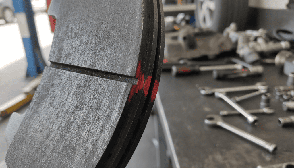 A close-up view of a brake wear indicator on a metallic brake pad, showcasing visible signs of wear. The brake pad has grooves and a slightly worn surface, with a bright red wear indicator line contrasting against the dark metallic colors. The foreground focuses on the brake pad, with a blurred background of an automotive mechanic's workshop, featuring tools and parts in a soft-focus. The lighting is bright and natural, mimicking daylight filtering through a workshop window, enhancing the details on the brake pad. The atmosphere is technical and informative, evoking a sense of urgency for maintenance. The angle is slightly tilted to emphasize the wear indicator clearly.